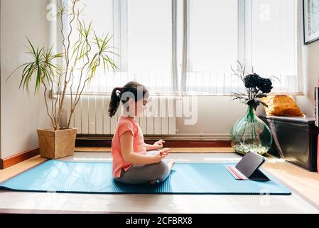 Kleines Mädchen, das in Lotuspose sitzt und auf einer Matte meditiert Mit geschlossenen Augen zu Hause mit großem Fenster beim Üben Yoga und Video-Tutorials auf dem Tablet Stockfoto