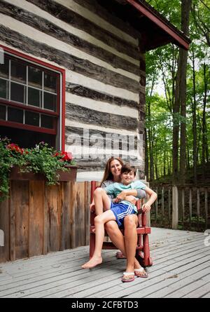 Mutter und Sohn sitzen im Schaukelstuhl auf Deck einer rustikalen Blockhütte. Stockfoto