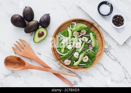 Flaches Lay von runden Holzteller mit Salat von Grün Und Avocado garniert mit roter Zwiebel und Garnelen mit Zutaten auf weißem Marmor Stockfoto