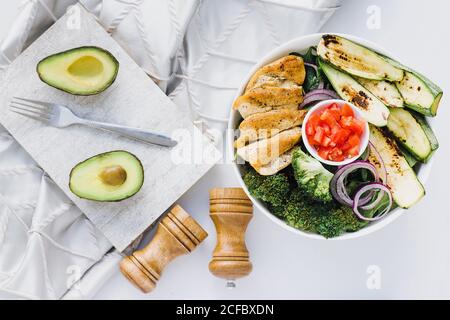 Von oben gehackte Tomaten in eine Schüssel mit gebratenem Huhn gelegt Und Zucchini mit Brokkoli und roten Zwiebeln in der Nähe halbierte Avocado Und Gewürze Stockfoto