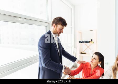 Super Arbeit! Zwei fröhliche Geschäftsleute, die sich die Hände schütteln, während ihre Kollegen im Hintergrund applaudieren und lächeln Stockfoto