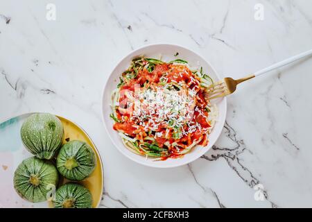 Von oben köstliche gesunde Mischung aus frischen grünen Nudeln hergestellt Von runden Zucchini mit Tomatensauce und gekrönt mit geriebenen Käseplatte auf Marmoroberfläche mit Gabel Stockfoto