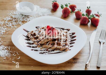 Weiche, süße Waffeln und Erdbeeren auf dem Teller mit leckeren Speisen Schokoladensirup und Kokosflocken auf Holztisch neben Besteck Stockfoto