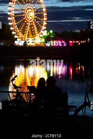 München, Deutschland. September 2020. Ein Mann und eine Frau sitzen im Olympiapark an einem Teich, in dem bunte Lichter von verschiedenen Fahrgeschäften reflektiert werden. Die Fahrgeschäfte und andere Attraktionen wurden eingerichtet, um die Showmen zu unterstützen. Durch die diesjährige Absage des Oktoberfestes haben viele Unternehmen finanzielle Verluste erlitten. Quelle: Sven Hoppe/dpa/Alamy Live News Stockfoto