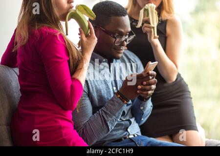 Dreiecksbeziehung. Zwei Frauen schauen auf schwarzen Mann und essen Banane, während Männer zwischen ihnen sitzen und Telefon Stockfoto