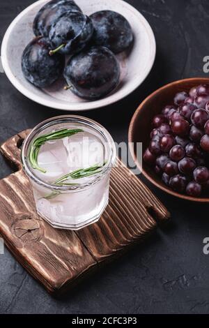 Frisches, eiskaltes kohlensäurehaltiges Wasser im Glas auf Schneidebrett mit Rosmarinblatt in der Nähe von Holzschalen mit Trauben- und Pflaumenfrüchten, dunkler Steinhintergrund Stockfoto