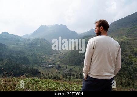 Rückansicht des Reisenden männlichen im lässigen Outfit stehend auf Grüner Hügel und genießen Sie einen schönen Blick auf Berggipfel während Nebliger Morgen Stockfoto