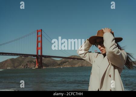 Seitenansicht einer jungen Frau in trendigem Outfit mit Hut und Sonnenbrille, die an sonnigen Tagen auf dem Damm gegen die Golden Gate Bridge in Kalifornien steht Stockfoto