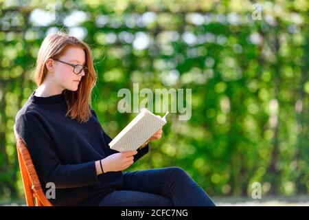 Mädchen sitzt im Frühling auf einem Stuhl im Garten Und liest in einem Buch Stockfoto