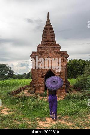 Rückansicht der Frau in langen Kleid hält traditionelle Burma Lila Regenschirm, während auf grüner Wiese vor stehen Alter Steintempel in der alten Stadt Bagan in bewölktem Tag Stockfoto