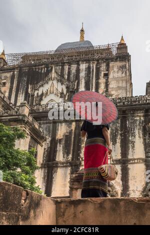 Von unten Rückansicht einer Frau in langen Kleidern, die einen traditionellen birmanischen roten Regenschirm und eine Tasche in der Nähe eines alten Steingebäudes hält, während sie in Bagan Myanmar spazieren und Sehenswürdigkeiten besichtigen Stockfoto