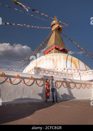 Paar hält sich die Hände und schaut einander im Stehen an In der Nähe buddhistischer Tempel mit dekorativen Girlanden und Turm unter Wolken Himmel bei Tageslicht Stockfoto