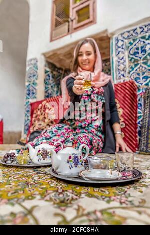 Lächelnde junge iranische Frau in traditionellem farbenfrohem Kleid und Schal auf dem Kopf sitzend auf dem mit Teppich bedeckten Boden vor Tabletts mit schönem Teeset und Glas mit Tee in ausgestreckter Hand haltend Stockfoto