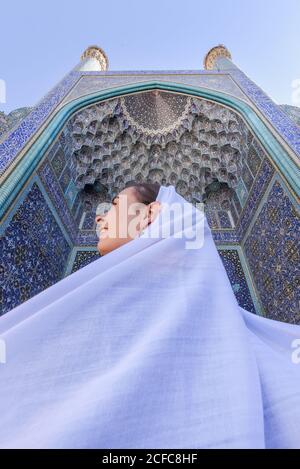 Von unten sehen Sie eine lächelnde junge muslimische Frau in Flieder-Hijab, die an sonnigen Tagen vor einer wunderschönen Moschee mit Mosaikwänden im Iran steht Stockfoto