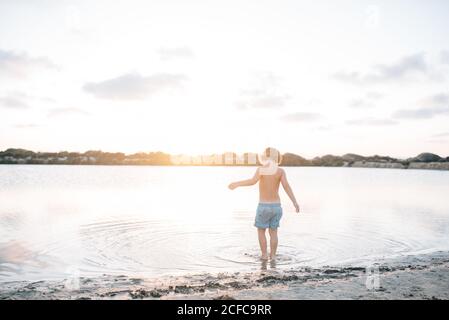 Rückansicht des verträumten Jungen, der im seichten Wasser läuft Strand gegen Sonnenuntergang Licht Stockfoto