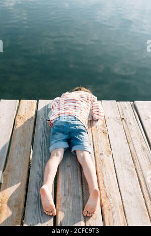 Von oben schaukelt das Kind mit dem Gesicht nach unten Hölzerner Pier in Sonnenschein mit friedliches Wasser unten Stockfoto