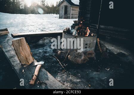 Brennendes Lagerfeuer und Log mit Axt in der Nähe einer kleinen Hütte platziert Von Holzfäller in verschneiten Wald im Winter Tag auf dem Land Von Finnland Stockfoto