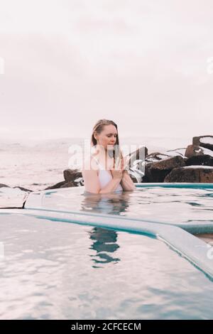 Junge Frau mit geschlossenen Augen, die sich im Wasser des Pools in der Nähe von Felsen und bewölktem Himmel ruhte Stockfoto