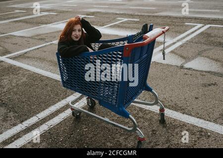Attraktive junge Frau mit roten Haaren in schwarzer Jacke, die Spaß hat, im Warenkorb auf dem markierten Parkplatz zu sitzen Stockfoto
