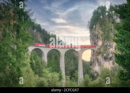 Malerischer Blick auf den roten Zug, der an der hohen Bogenbrücke vorbeifährt Tunnel in den Bergen während der Fahrt durch grünen Wald in der Schweiz Alpen Stockfoto