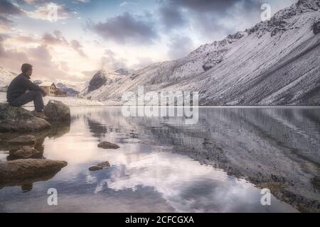 Seitenansicht eines erwachsenen Mannes, der auf Stein sitzt, nahe der Ruhe see und schneebedeckten Berg und bewundern wolkigen Sonnenuntergang Himmel während Besuch des Schweizer Nationalparks in der Schweiz Stockfoto