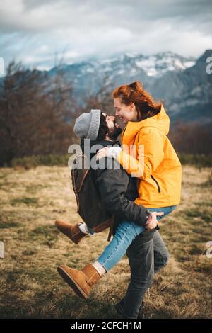 Seitenansicht eines amourösen Mannes mit Rucksack, der eine lächelnde Frau in der Hand hält und sich am bewölkten Tag mit trockenem Gras in der Nähe der Berge amüsiert Stockfoto