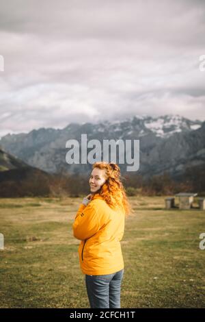Rückansicht einer lächelnden Frau in einer warmen gelben Jacke, die die Kamera über der Schulter auf einer grünen Wiese, umgeben von verschneiten Bergen, anschaut Stockfoto