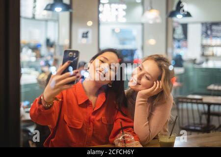 Zwei junge Freundinnen mit Tassen frischen Kaffee lächeln und posieren für Selfie, während sie am Tisch in einem gemütlichen Restaurant sitzen und ein Getränk trinken Stockfoto