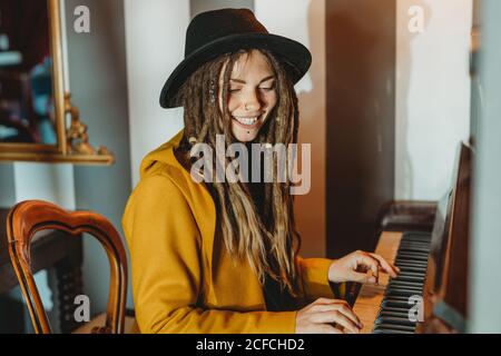 Seitenansicht einer glücklichen Hipster-Hündin mit Dreadlocks in Gelb Mantel und schwarzer Hut spielen Klavier, während im Retro sitzen Zimmer im Stil Stockfoto