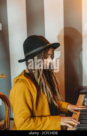 Seitenansicht einer ernsthaften Hipster-Hündin mit Dreadlocks in Gelb Mantel und schwarzer Hut spielen Klavier, während im Retro sitzen Zimmer im Stil Stockfoto