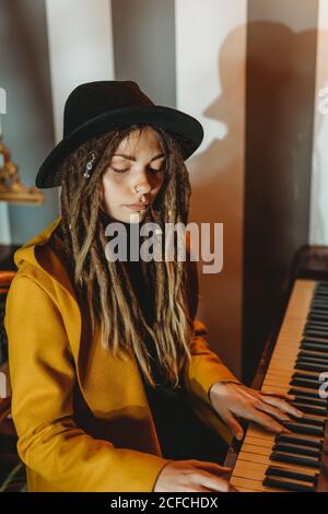 Seitenansicht einer ernsthaften Hipster-Hündin mit Dreadlocks in Gelb Mantel und schwarzer Hut spielen Klavier, während im Retro sitzen Zimmer im Stil Stockfoto