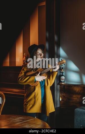 Hipster Millennial Weibchen mit Dreadlocks in gelbem Mantel und schwarz hut spielt Hawaiian Gitarre Ukulele, während im dunklen vintage stehen Zimmer Stockfoto