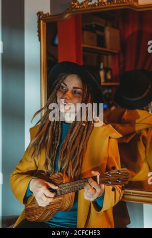 Junge stilvolle Frau mit Dreadlocks in gelbem Mantel und schwarzem Hut, die auf einem alten Holztisch sitzt, zurück zum Spiegel und Hawaii-Gitarren-Ukulele im Zimmer mit antiken Möbeln spielt Stockfoto