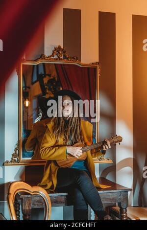 Junge stilvolle Frau mit Dreadlocks in gelbem Mantel und schwarzem Hut, die auf einem alten Holztisch sitzt, zurück zum Spiegel und Hawaii-Gitarren-Ukulele im Zimmer mit antiken Möbeln spielt Stockfoto