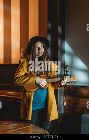 Hipster Millennial Weibchen mit Dreadlocks in gelbem Mantel und schwarz hut spielt Hawaiian Gitarre Ukulele, während im dunklen vintage stehen Zimmer Stockfoto