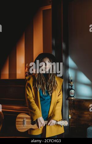 Hipster Millennial Weibchen mit Dreadlocks in gelbem Mantel und schwarz hut spielt Hawaiian Gitarre Ukulele, während im dunklen vintage stehen Zimmer Stockfoto