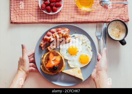 Anonyme Ernte Hände Putting Teller mit leckeren Spiegeleiern mit Speck und Käse auf dem Tisch zum Frühstück Stockfoto