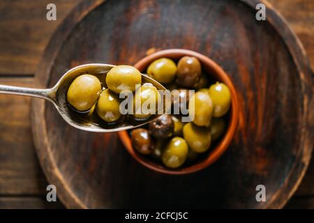 Draufsicht auf Metalllöffel mit Oliven in Öl auf Hintergrund der Keramikschale auf Holztisch in der Küche platziert Stockfoto