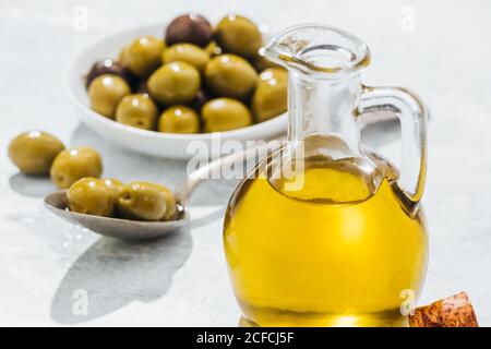 Glaskrug mit Öl und kleine Schüssel und Löffel mit Oliven auf weißem Tisch Stockfoto