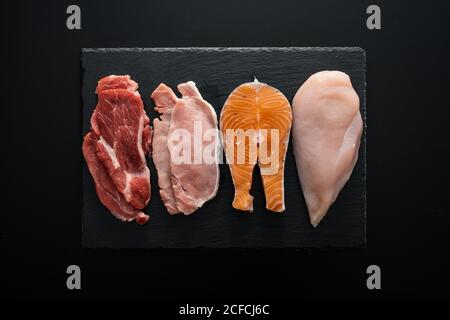 Von oben frisches rohes Rinderfilet und Schweinefleisch mit Huhn und Fisch Stücke auf Holz Schneidebrett als Zusammensetzung Von eiweißreichen Lebensmitteln vor schwarzem Hintergrund Stockfoto