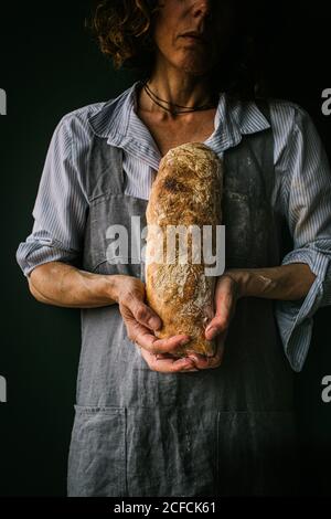 Eine nicht erkennbare Frau auf der Schürze, die Ciabatta-Brot hält, während sie auf dunklem Hintergrund steht Stockfoto
