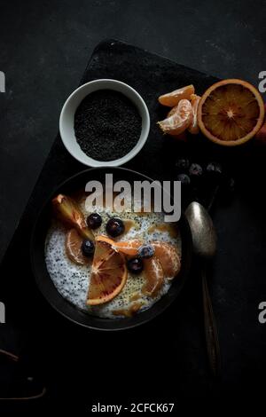 Von oben Chia Samen Pudding mit Scheiben von Orange, Mandarinen und Oliven in Schüssel neben Löffel und Grapefruit auf dunklem Tisch Stockfoto