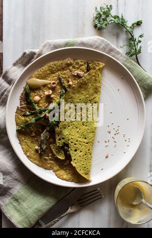 Draufsicht auf weißen Teller mit Haferkrepe mit Spargel Und Tahini-Paste serviert auf rustikalem Brett mit Grüns Stockfoto