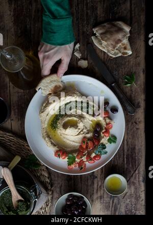 Von der oberen Hand der anonymen Person tauchend Stück frisch Fladenbrot in leckeres Hummus Pesto mit Kirschtomaten und dekoriert Bohnen mit Petersilie über Holzplatte Stockfoto