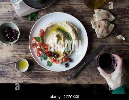 Von der oberen Hand der anonymen Person tauchend Stück frisch Fladenbrot in leckeres Hummus Pesto mit Kirschtomaten und dekoriert Bohnen mit Petersilie über Holzplatte Stockfoto