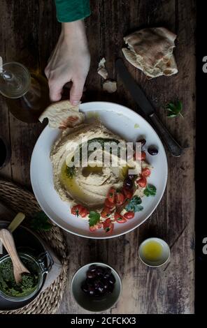 Von der oberen Hand der anonymen Person tauchend Stück frisch Fladenbrot in leckeres Hummus Pesto mit Kirschtomaten und dekoriert Bohnen mit Petersilie über Holzplatte Stockfoto