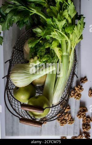 Von oben Metallkorb mit Äpfeln, Sellerie, Fenchelknollen und Walnüssen an Bord vor weißem Hintergrund angeordnet Stockfoto