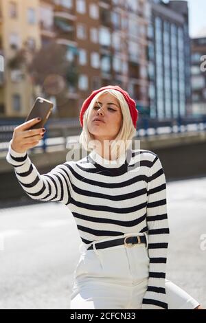 Junge lächelnde hübsche Frau mit roter französischer Mütze, gestreifter Bluse und weißen Shorts, die auf urbanem Hintergrund fotografiert Stockfoto