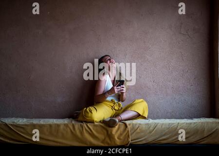 Fröhliche blonde Frau, die auf dem Sofa auf der Terrasse im orientalischen Stil sitzt und in Marokko ein Mobiltelefon benutzt Stockfoto