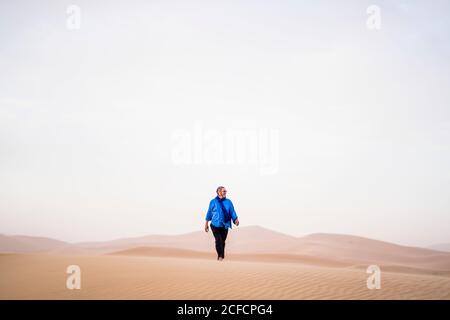Reifer stilvoller bärtiger Mann, der beim Spaziergang im Sand wegschaut Wüste gegen grauen Himmel in Marokko Stockfoto